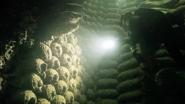 Photographie sous-marine montrant une structure submergée, possiblement une épave ou des ruines, éclairée par une lumière vive dans une eau trouble, illustrant les fonds marins où des centaines de milliers de tonnes d'armes conventionnelles et chimiques ont été immergées au large des côtes française