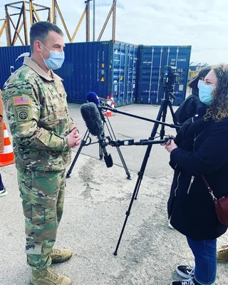 Le lieutenant-colonel Jake Whiteside de l'armée américaine, en uniforme de camouflage et masque facial bleu, répond aux questions d'une journaliste avec micro et caméra sur trépied, devant des conteneurs bleus au port de Dunkerque.