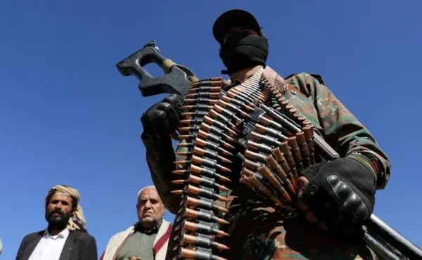 Un militant houthi masqué, vêtu de camouflage et chargé de ceintures de munitions, tient une mitrailleuse devant deux hommes plus âgés en tenue traditionnelle sous un ciel bleu, dans un article sur la puissance des dirigeants rebelles du Yémen.