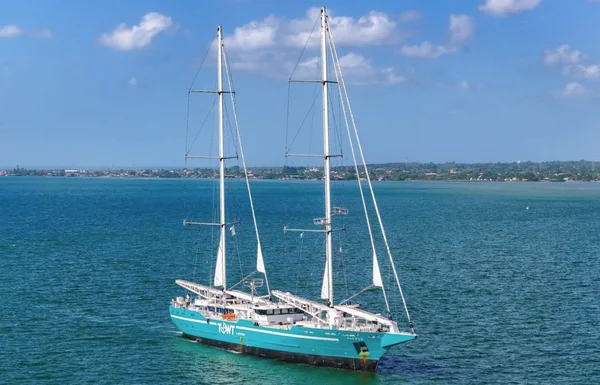 Le voilier TOWT, compagnie française de transport à la voile pour la transition maritime, à coque teal et deux mâts, flottant sur eau bleue calme avec littoral lointain et ciel nuageux.