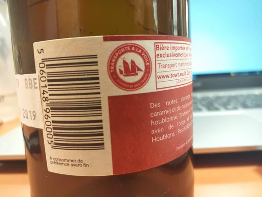 Gros plan sur l'étiquette arrière d'une bouteille de bière en verre brun, montrant un code-barres, du texte français et le logo 'Transporté à la voile' de TOWT pour ses premières marchandises expédiées à la voile.