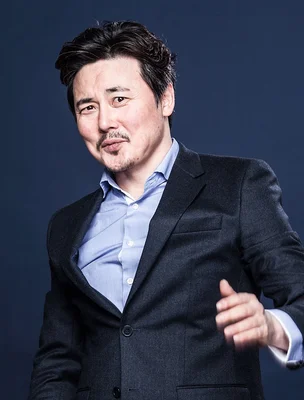 Portrait en studio de Na Jae-Woong, homme d'âge moyen aux cheveux sombres et à la moustache, portant une veste sombre sur une chemise bleu clair, souriant légèrement à la caméra.