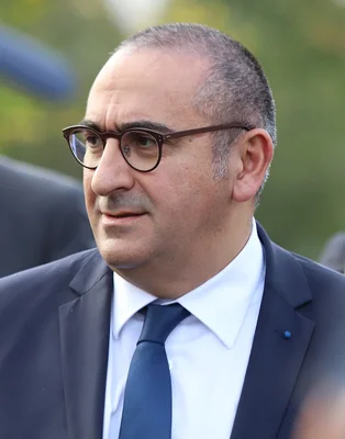 Laurent Nuñez, homme politique, en gros plan avec cheveux courts grisonnants et lunettes, vêtu d'un costume sombre, chemise blanche et cravate bleue, regardant légèrement vers la droite.