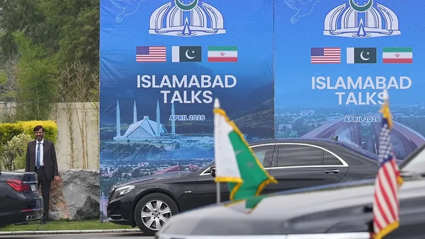 Photographie extérieure des 'Islamabad Talks' d'avril 2026 : bannières avec drapeaux des États-Unis, du Pakistan et d'Iran, un homme en costume à gauche, voitures pavoisées, évoquant les négociations tendues.