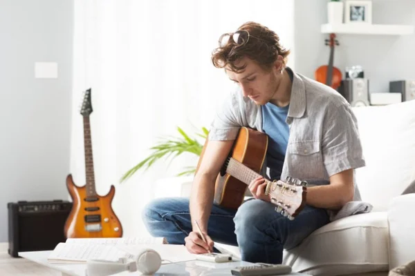 Jeune homme aux cheveux bouclés assis sur un canapé blanc tenant une guitare acoustique et écrivant sur une feuille sur une table basse, illustrant la profession de parolier, avec guitare électrique et ampli en arrière-plan.