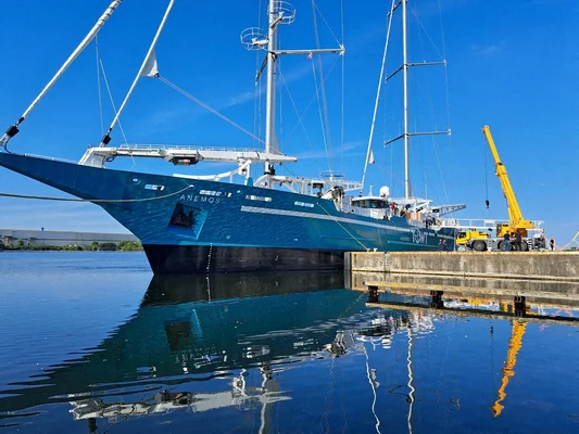 Anemos, le premier voilier cargo de la compagnie du Havre, un grand yacht à voile bleu nommé ANEMOS amarré à un quai en béton sous un ciel bleu dégagé, avec sa réflexion dans l'eau calme et une grue jaune à proximité.