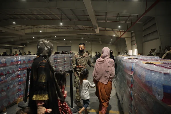 Le sergent Juan Miranda, spécialiste culinaire du 155e bataillon de soutien au combat, en uniforme de camouflage et masque noir, accueille une famille de femmes et enfants afghans dans un entrepôt du camp As Sayliyah au Qatar, le 20 août 2021, près de palettes d'eau.