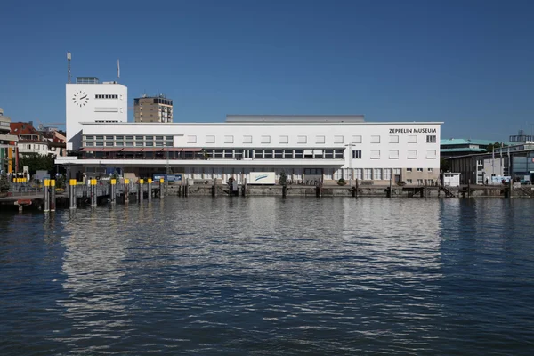 Le Musée Zeppelin, grand bâtiment blanc avec une tour horloge, situé en bord de plan d'eau sous un ciel bleu dégagé.