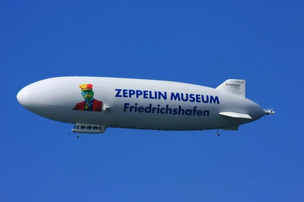 Un dirigeable Zeppelin NT blanc arborant l'inscription 'ZEPPELIN MUSEUM Friedrichshafen' volant dans un ciel bleu clair, symbole du musée de Friedrichshafen.