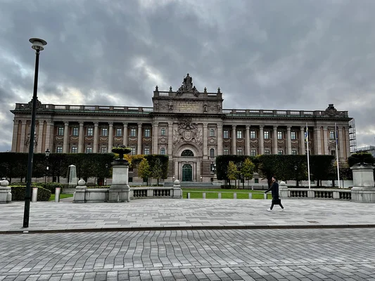 Le Parlement de Suède (Riksdag) à Gamla Stan, Stockholm, sous un ciel nuageux, avec une personne traversant la place pavée au premier plan.