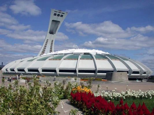 Le Stade olympique de Montréal, avec sa tour inclinée et son toit blanc en dôme, des parterres de fleurs colorés à l'avant-plan sous un ciel bleu.