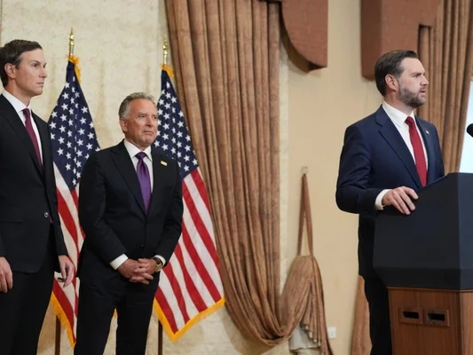 Trois hommes en costume dans une salle officielle avec drapeaux américains, l'un parlant au podium et les deux autres derrière lui, lors des négociations États-Unis-Iran avec le Pakistan comme médiateur.