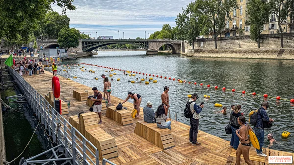 Sur une plateforme flottante en bois sur la Seine à Paris, des personnes nagent et se détendent sous un pont de pierre et des immeubles parisiens, dans le cadre des sites de baignade gratuits en Île-de-France (Sortiraparis.com).