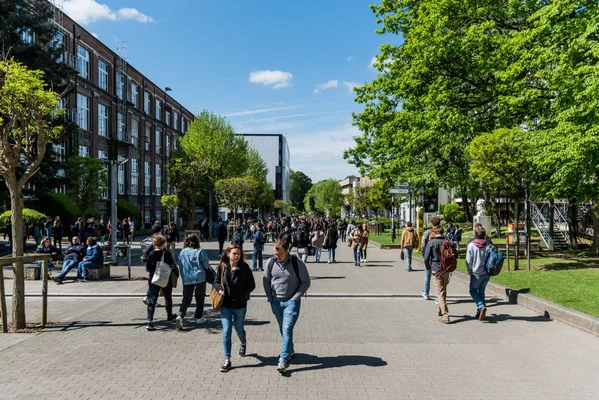 Campus de l'université libre de Bruxelles, vue large d'une allée piétonne animée par temps ensoleillé avec des gens marchant et assis près d'un bâtiment en brique et d'arbres.