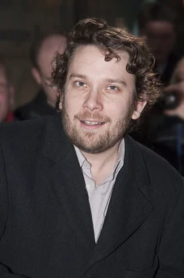 Christian Ulmen, vu de plus près, avec des cheveux bruns bouclés et une barbe, portant une veste sombre sur une chemise claire à rayures, image issue de Wikipédia.