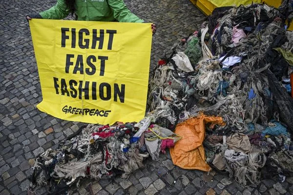 Bientôt une taxe sur la fast-fashion, catastrophe sociale et écologique ? Manifestant en veste verte tenant banderole jaune Greenpeace 'FIGHT FAST FASHION' près de tas de vêtements sur pavés.