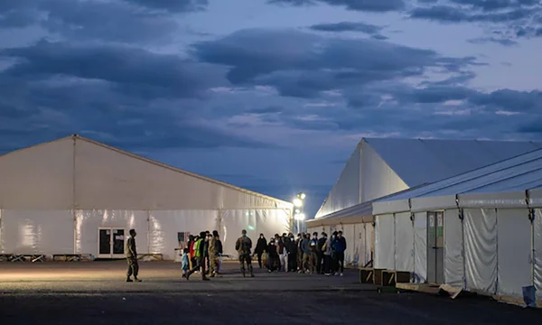 Des personnes, dont des militaires, marchent entre de grandes tentes blanches dans un camp d'évacués afghans au Qatar au crépuscule, alors que Trump veut le fermer, suscitant débat.