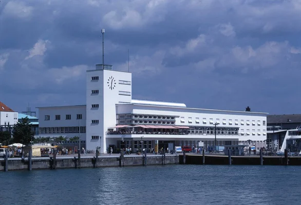 Le Musée Zeppelin de Friedrichshafen, bâtiment blanc moderniste à tour horloge, avec aile horizontale et terrasse, en bord de lac sous ciel nuageux et ponton au premier plan.
