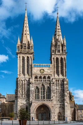 La cathédrale Saint-Paul-Aurélien de Saint-Pol-de-Léon, façade gothique en pierre claire avec deux flèches pointues et horloge, ornée de sculptures, sous un ciel bleu nuageux.