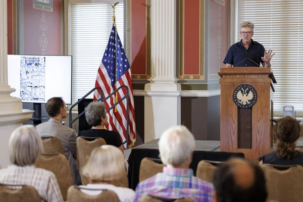 Derf Backderf, auteur et artiste récompensé, aux cheveux gris courts et lunettes, parlant à un podium en bois devant un public, avec drapeau américain et écran affichant une bande dessinée, lors d'une conférence sur son roman graphique Kent State.