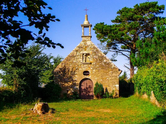 La chapelle et le colombier de Kérigou à Saint-Pol-de-Léon, petite bâtisse rustique en pierre avec clocheton et croix, entourée d'herbe et d'arbres sous un ciel bleu.