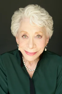 Bunny Levine, femme âgée aux cheveux courts et ondulés blancs, portant un chemisier vert foncé et un collier de perles, souriant à l'appareil photo sur fond sombre.