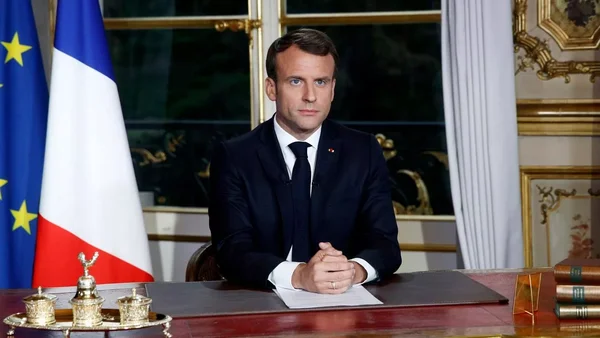 Le président de la République en costume sombre et cravate, assis à un bureau aux mains jointes devant les drapeaux français et européen dans une salle ornée.