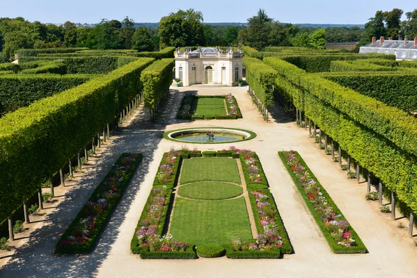 Jardin français du Petit Trianon au Château de Versailles : parterres de fleurs symétriques, haies manicurées, fontaine centrale et pavillon classique en arrière-plan, lieu évoquant un temps suspendu.