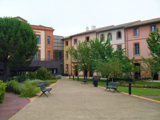 Centre Hospitalier de Muret : allée pavée dans une cour entourée de bâtiments à façades colorées et d'espaces verts, avec bancs modernes sous un ciel couvert.