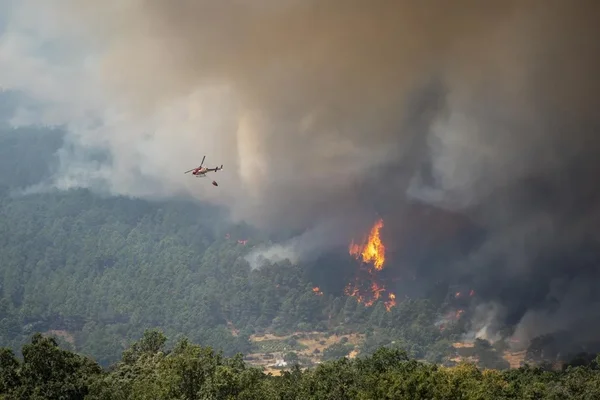 Espagne : état d'urgence nationale face aux méga-feux. Un hélicoptère de pompiers larguant de l'eau sur un grand incendie en pente boisée avec d'épaisses fumées.