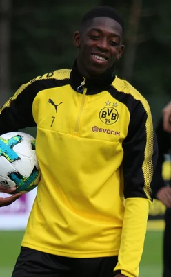 Ousmane Dembélé, souriant, portant un maillot d'entraînement jaune et noir du Borussia Dortmund et tenant un ballon de football sous le bras droit, lors d'un stage à Bad Ragaz en 2017.