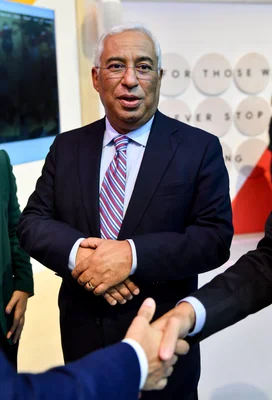 António Costa, alors Premier ministre du Portugal, homme aux cheveux blancs et portant des lunettes, en costume sombre et cravate à rayures, les mains jointes lors du Web Summit 2019 à Lisbonne, avec une poignée de main au premier plan.