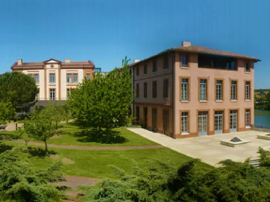 Centre Hospitalier de Muret : grand bâtiment de plusieurs étages à façade rouge-brun avec de nombreuses fenêtres, près d'un plan d'eau, entouré de pelouses et arbres sous ciel bleu.