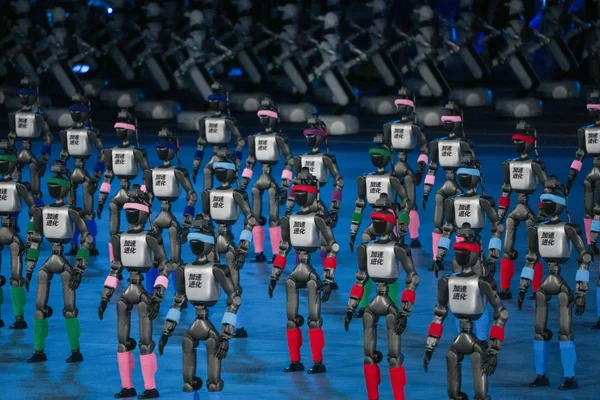 Cérémonie d'ouverture des deuxièmes Jeux mondiaux des robots humanoïdes à Beijing : un grand groupe de robots avec bandeaux colorés et guêtres exécutent une routine synchronisée sur scène bleue, panneau ' ' sur la poitrine