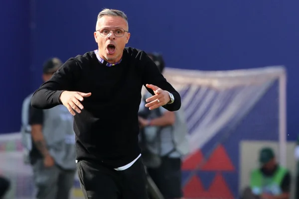 Mohamed Ouahbi, ancien prof de sport et sélectionneur des Lions de l'Atlas, cheveux courts gris et lunettes, portant un pull noir, criant et gesticulant sur le bord du terrain de football.