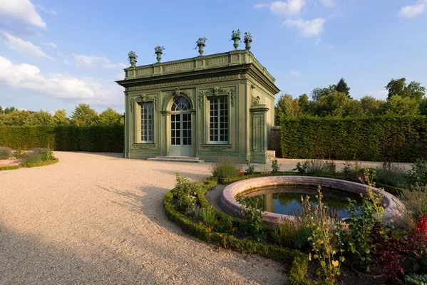 Pavillon de jardin vert orné du Petit Trianon à Versailles, grandes fenêtres et urnes décoratives sur le toit, chemin de gravier et fontaine circulaire en premier plan, espace intemporel.