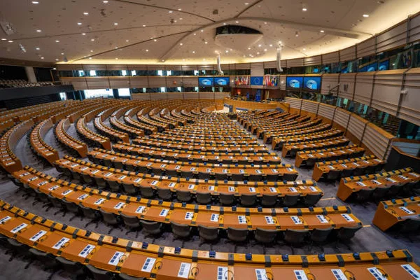 Salle plénière de l’hémicycle du Parlement européen à Bruxelles lors d'une séance plénière, chambre vide avec rangées de bureaux et chaises en bois courbes en demi-cercle, podium central avec écrans et drapeaux sous un plafond haut.