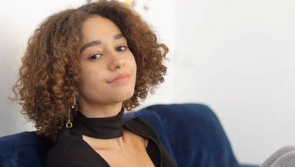 Léna Situations, influenceuse, assise sur un canapé bleu, cheveux bouclés bruns et haut noir, regardant la caméra avec un léger sourire, dans un reportage sur ses ventes de livres dépassant Marc Levy et Guillaume Musso.
