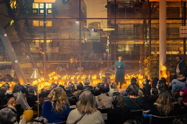 Vue du théâtre en version immersive, de près ou de loin : une femme en robe bleue sur scène parmi des bougies scintillantes s'adresse à un public dans une salle moderne aux parois vitrées la nuit.