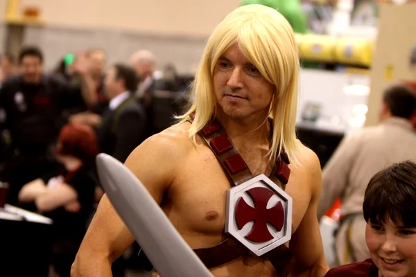 Cosplayeur habillé en He-Man avec perruque blonde et harnais pectoral à croix de fer, debout dans un hall de convention lors de la Phoenix Comicon 2012 à Phoenix, Arizona.