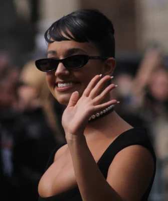 Léna Situation arrivant au défilé MiuMiu Femme Printemps/Été 2025, cheveux sombres en chignon avec frange courte, lunettes de soleil noires et haut noir, souriant et faisant un signe de la main vers la caméra.