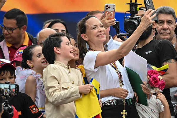 María Corina Machado à Ciudad de Panamá, portant une chemise blanche, prenant un selfie avec des enfants et adultes souriants tenant des drapeaux vénézuéliens dans un lieu extérieur bondé.