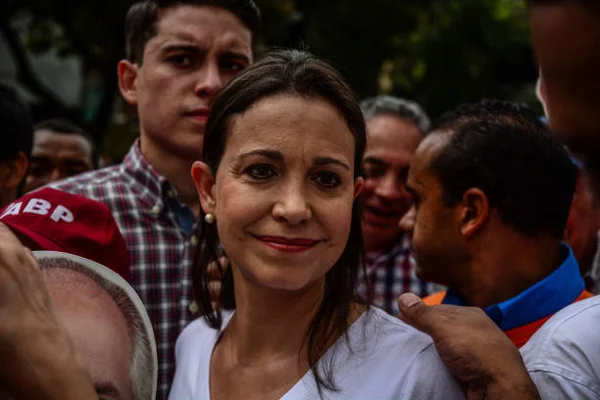 Maria Corina Machado, leader politique, lors d'une manifestation contre le gouvernement de Nicolas Maduro, souriant légèrement, cheveux foncés et lèvres rouges, entourée d'hommes en extérieur.