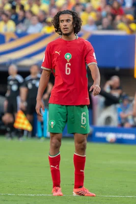 Ayyoub Bouaddi, joueur de l'équipe nationale du Maroc, portant le maillot rouge et vert numéro 6, debout sur un terrain d'herbe lors du match Brésil contre Maroc à la Coupe du Monde FIFA 2026 au stade NY/NJ à East Rutherford.