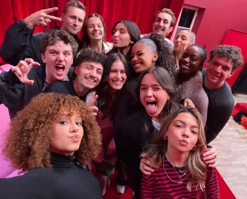 Léna Situations avec un groupe de jeunes adultes posant pour un selfie, faisant des grimaces et gestes ludiques devant un fond de rideau rouge, dans le contexte de sa réticence à venir au château de la Star Academy.