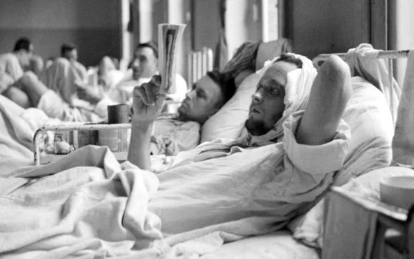 Photographie en noir et blanc de l'article "D'où vient l'hôpital ? On plonge dans l'histoire..." montrant un homme à la tête bandée alité lisant un journal, avec d'autres patients en arrière-plan.