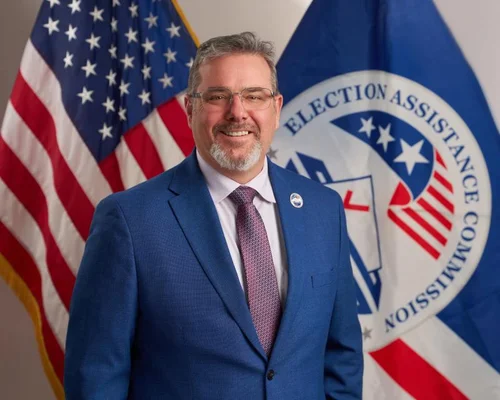 Donald Palmer, commissaire de la Commission d'aide aux élections (EAC) nommé par Trump en 2019, homme d'âge moyen avec lunettes et bouc, en costume bleu et cravate pourpre, souriant devant le drapeau américain et celui de l'EAC.