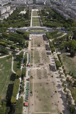 Vue en hauteur du Champ-de-Mars à Paris, montrant de longues pelouses vertes et des allées remplies de personnes, avec l'École Militaire au loin.