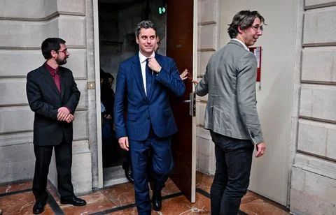 Gabriel Attal, adoubé par Renaissance pour 2027, en costume au centre d'un couloir en pierre ajustant sa cravate, accompagné de deux autres hommes en costume dont l'un ouvre une porte.