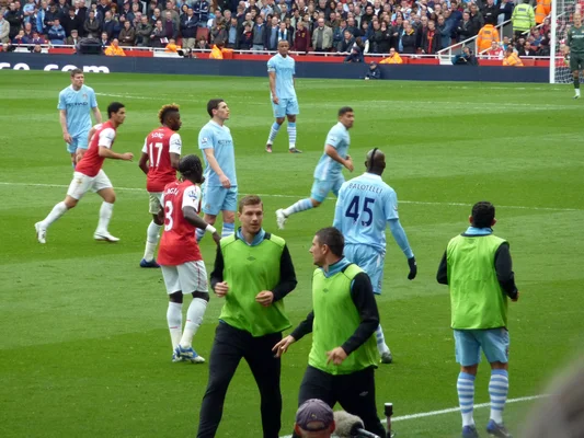 Joueurs de l'Arsenal et de Manchester City s'affrontant sur une pelouse verte, en maillots rouge et bleu clair, avec la foule en arrière-plan lors d'un match professionnel.